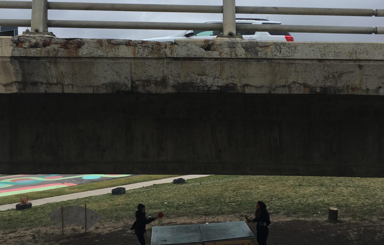 Two people playing ping pong under an elevated highway